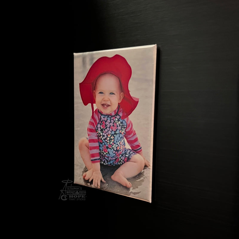 Close-up of a custom 2x3 photo magnet featuring a baby portrait displayed on a dark metal surface.