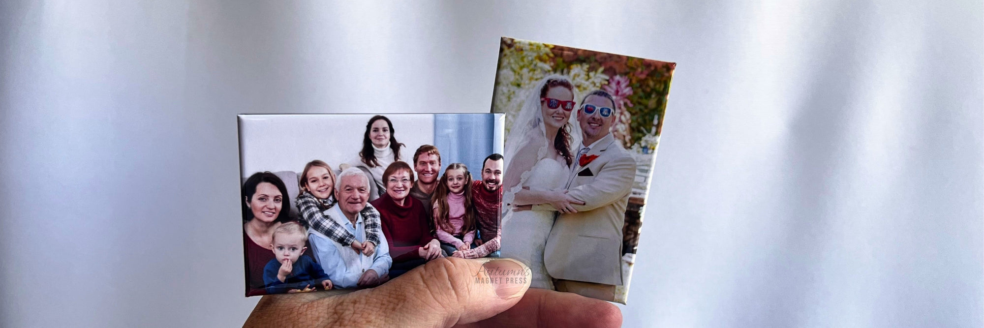 A hand holds two small custom photo prints against a soft white draped background. One print shows a group of people, and the other shows two women outdoors.