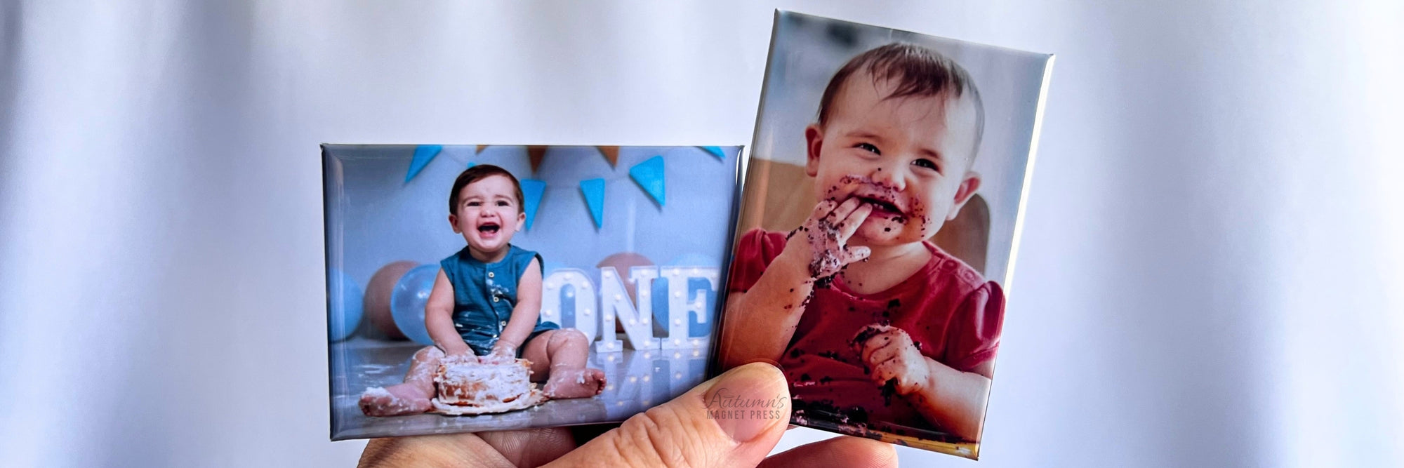 A hand holding two small custom photo magnets. One magnet shows a baby sitting on a floor with a birthday cake, and the other shows a smiling baby with chocolate smeared on their face and hands.