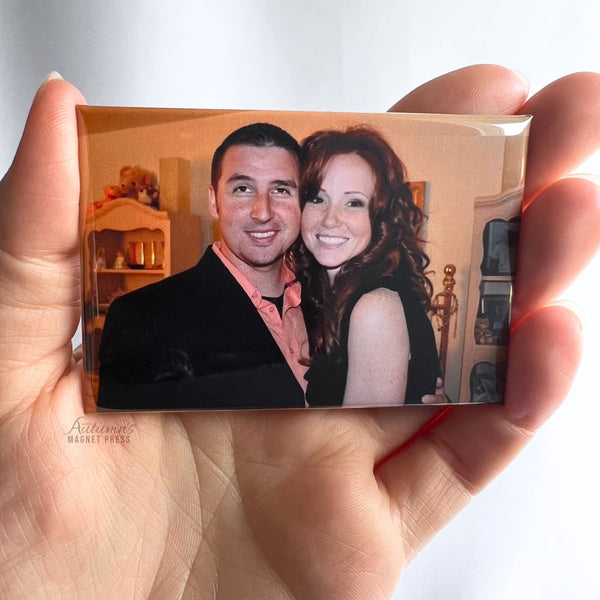 Hand holding a custom photo magnet featuring a smiling couple posing together to celebrate their anniversary against a soft white background.