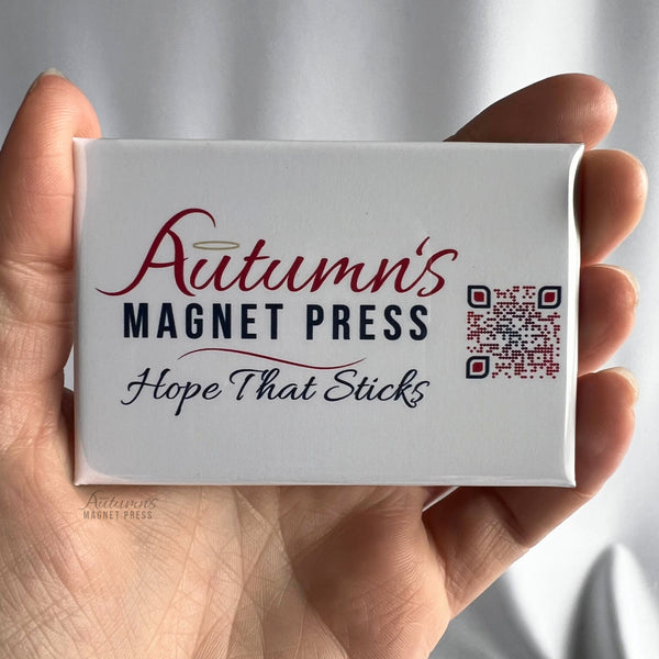 A hand holding a custom rectangular business logo magnet featuring the Autumn's Magnet Press logo printed on a white background. The design includes the name Autumn's Magnet Press in red and navy lettering, the tagline Hope That Sticks in elegant black script, and a QR code on the right side. The glossy personalized magnet showcases how businesses can turn their logos, branding, and contact information into promotional magnets for customers, events, and giveaways.