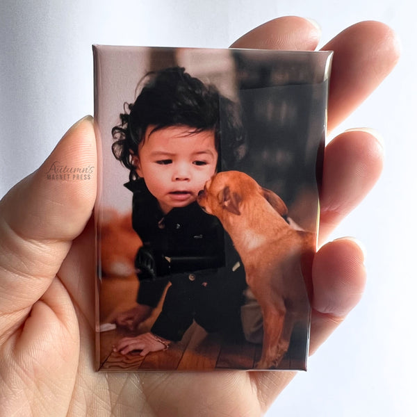 Hand holding a custom photo magnet featuring a young child sharing a moment with a dog against a soft white background.