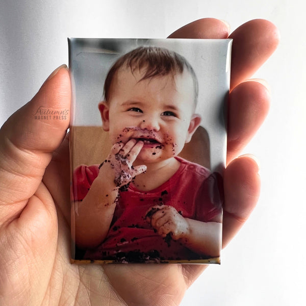 Hand holding a custom photo magnet featuring a smiling baby enjoying birthday cake during a first birthday celebration against a soft white background.