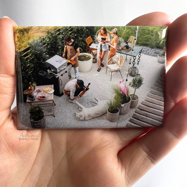 Hand holding a custom family photo magnet featuring loved ones gathered together in a backyard against a soft white background.
