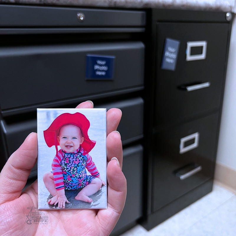 Custom baby photo magnet featuring a smiling baby in a red sun hat held in hand in front of black filing cabinets.