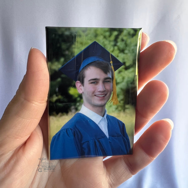 Hand holding a custom graduation portrait photo magnet featuring a young man in blue cap and gown outdoors against a soft white background