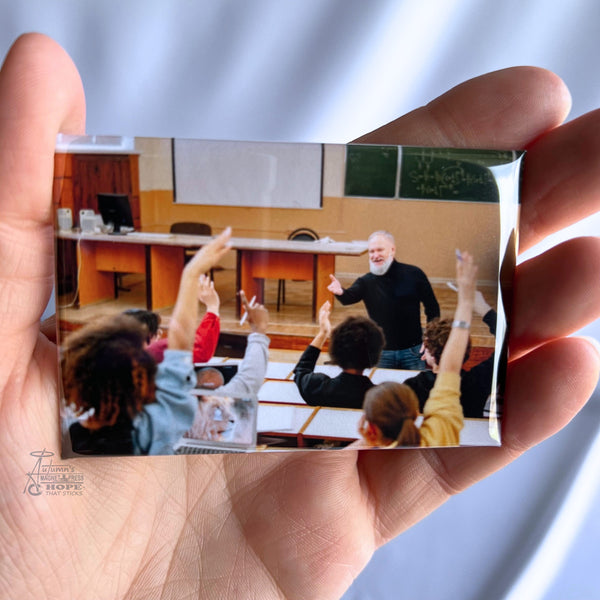 Hand holding a custom classroom photo magnet featuring a teacher speaking to students with raised hands against a soft white background