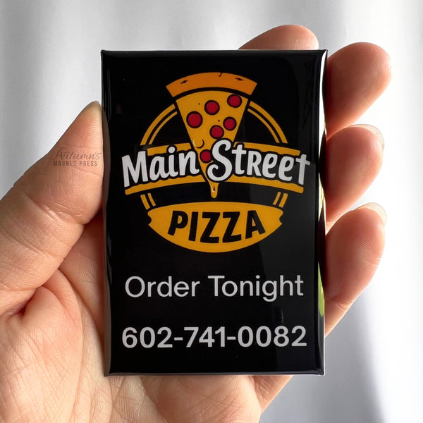 A hand holding a custom rectangular business promotional magnet featuring the Main Street Pizza logo with a colorful pizza slice, black and gold design, "Order Tonight" message, and business phone number 602-741-0082.