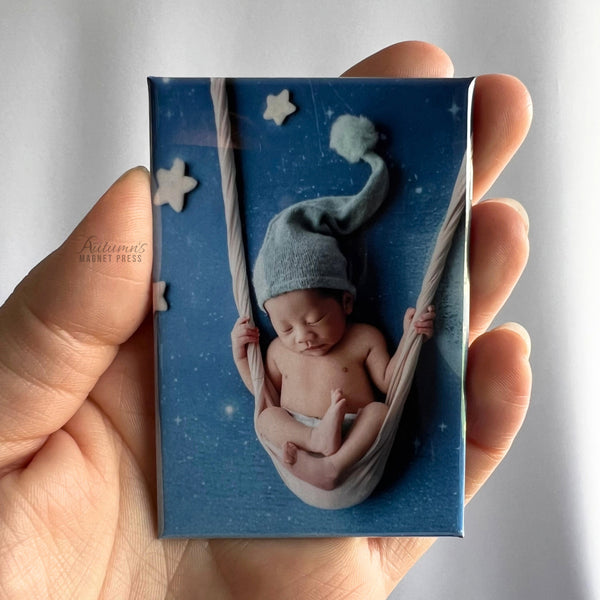 A hand holding a custom vertical rectangular newborn photo magnet featuring a sleeping baby wearing a gray knit hat, curled up in a crescent moon hammock suspended by ropes against a blue starry background with soft white clouds.