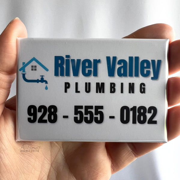 A hand holding a custom rectangular business advertising magnet featuring the River Valley Plumbing logo with a blue house, water faucet and droplet design, bold blue and black lettering, and the business phone number 928-555-0182 on a white background.