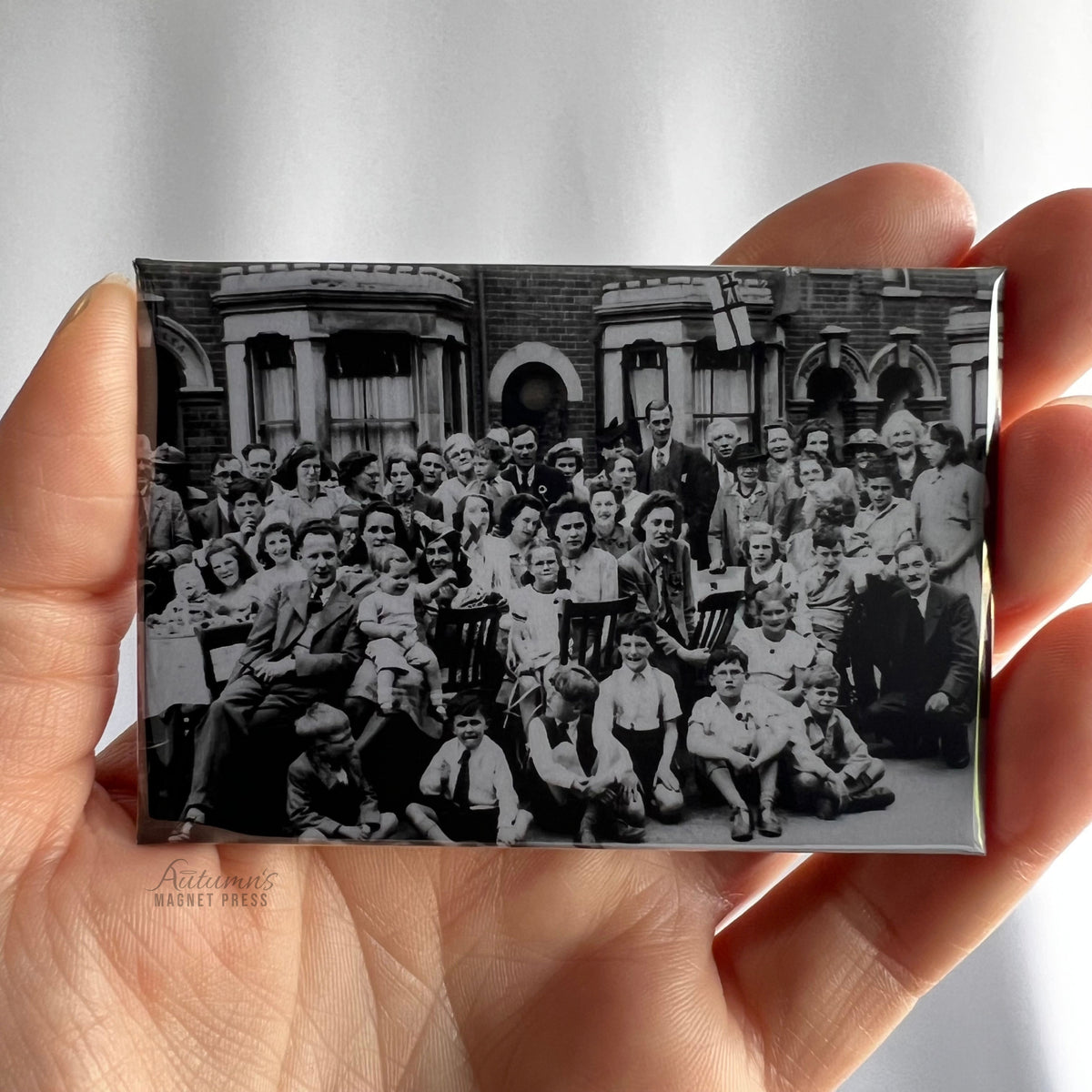 A hand holding a custom rectangular photo magnet featuring a vintage black-and-white group photograph of multiple generations of adults and children gathered together outside a brick building with arched windows, preserving a treasured family memory.
