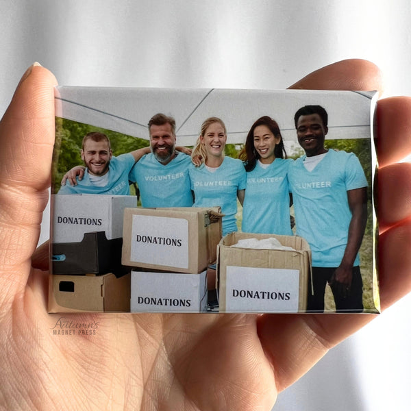 A hand holding a custom horizontal rectangular volunteer photo magnet featuring five smiling community volunteers wearing matching light blue shirts, standing together behind cardboard donation boxes under a white event tent during a charity donation drive.