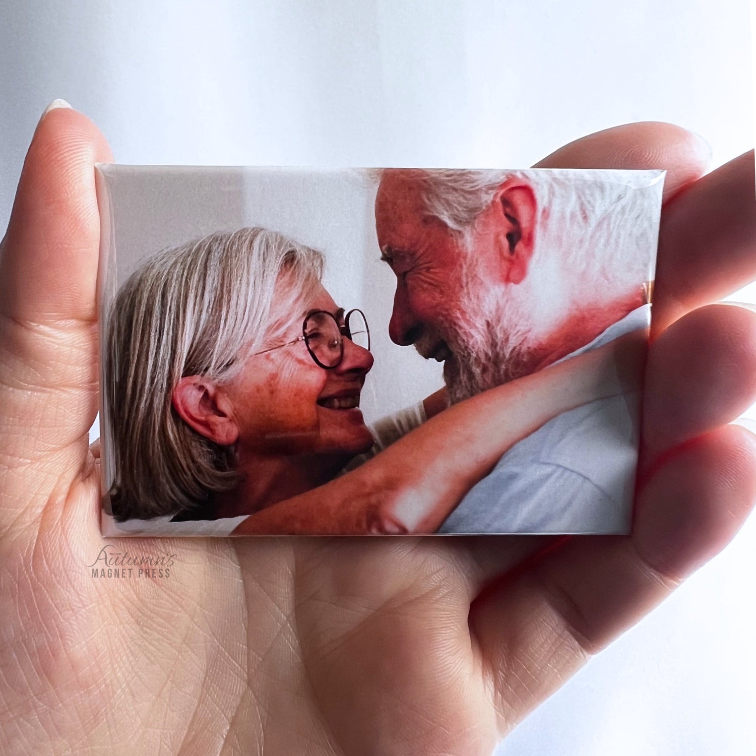 Hand holding a custom photo magnet featuring an elderly couple smiling at each other while embracing against a soft white background.