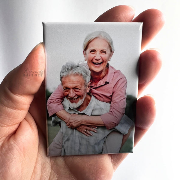 Hand holding a custom photo magnet featuring an elderly couple smiling and embracing outdoors against a soft white background