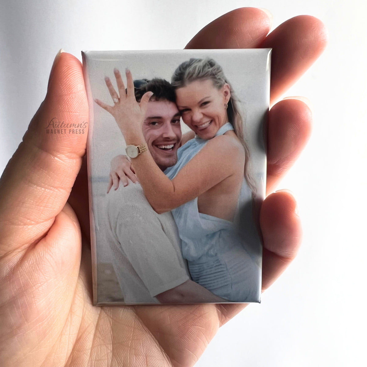 Hand holding a custom photo magnet featuring a smiling newly engaged couple embracing and showing an engagement ring against a soft white background.