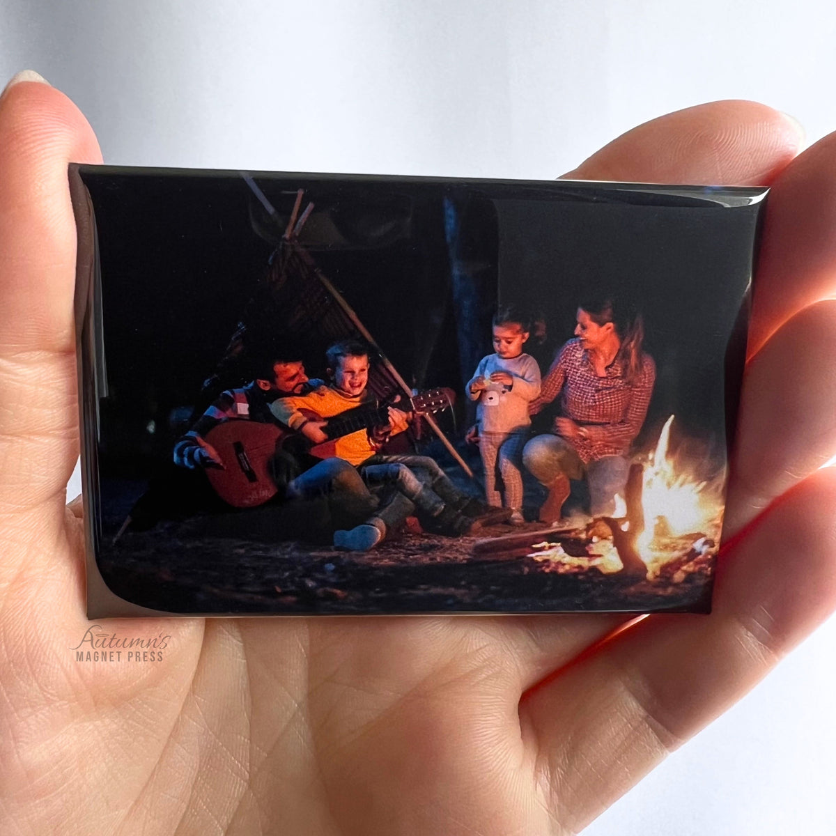 Hand holding a custom photo magnet featuring a family gathered around a campfire roasting marshmallows at night against a soft white background.