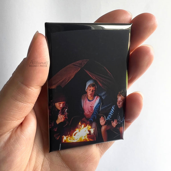 Hand holding a custom family photo magnet featuring loved ones sitting around a campfire during a camping trip against a soft white background.