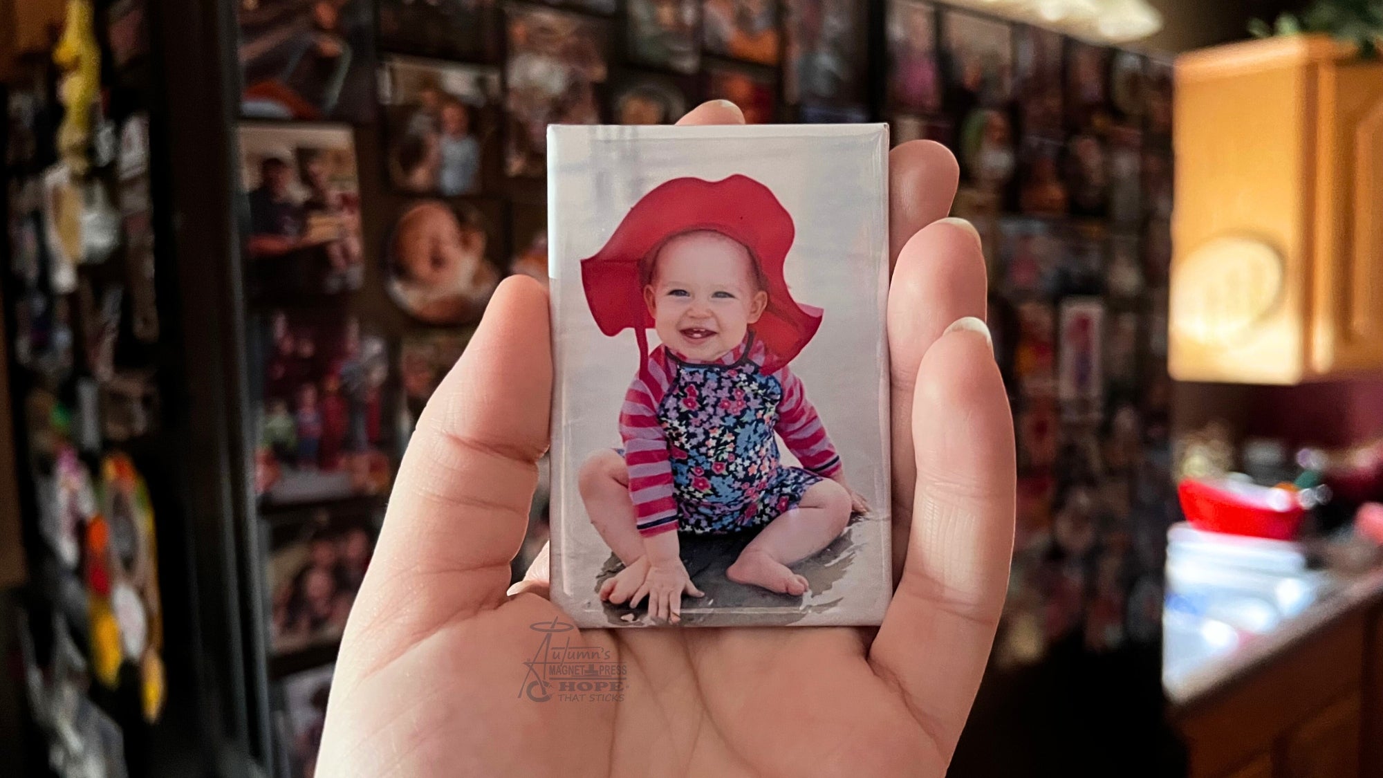 Hand holding a custom 2x3 family photo magnet featuring a smiling baby in a kitchen surrounded by photo-covered refrigerators and memory magnets.
