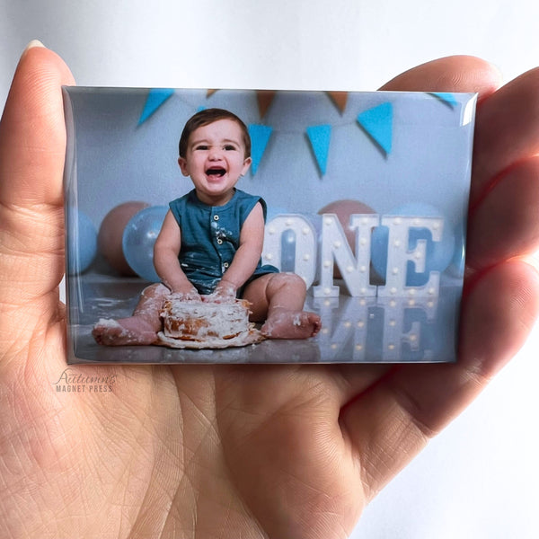 Hand holding a custom photo magnet featuring a smiling child celebrating a first birthday with a "ONE" sign and birthday cake against a soft white background.