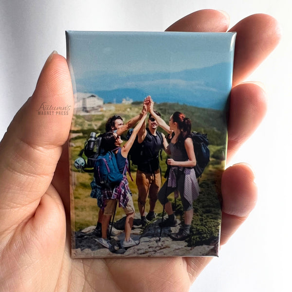 Hand holding a custom photo magnet featuring a group of friends celebrating together during a mountain hiking adventure against a soft white background.