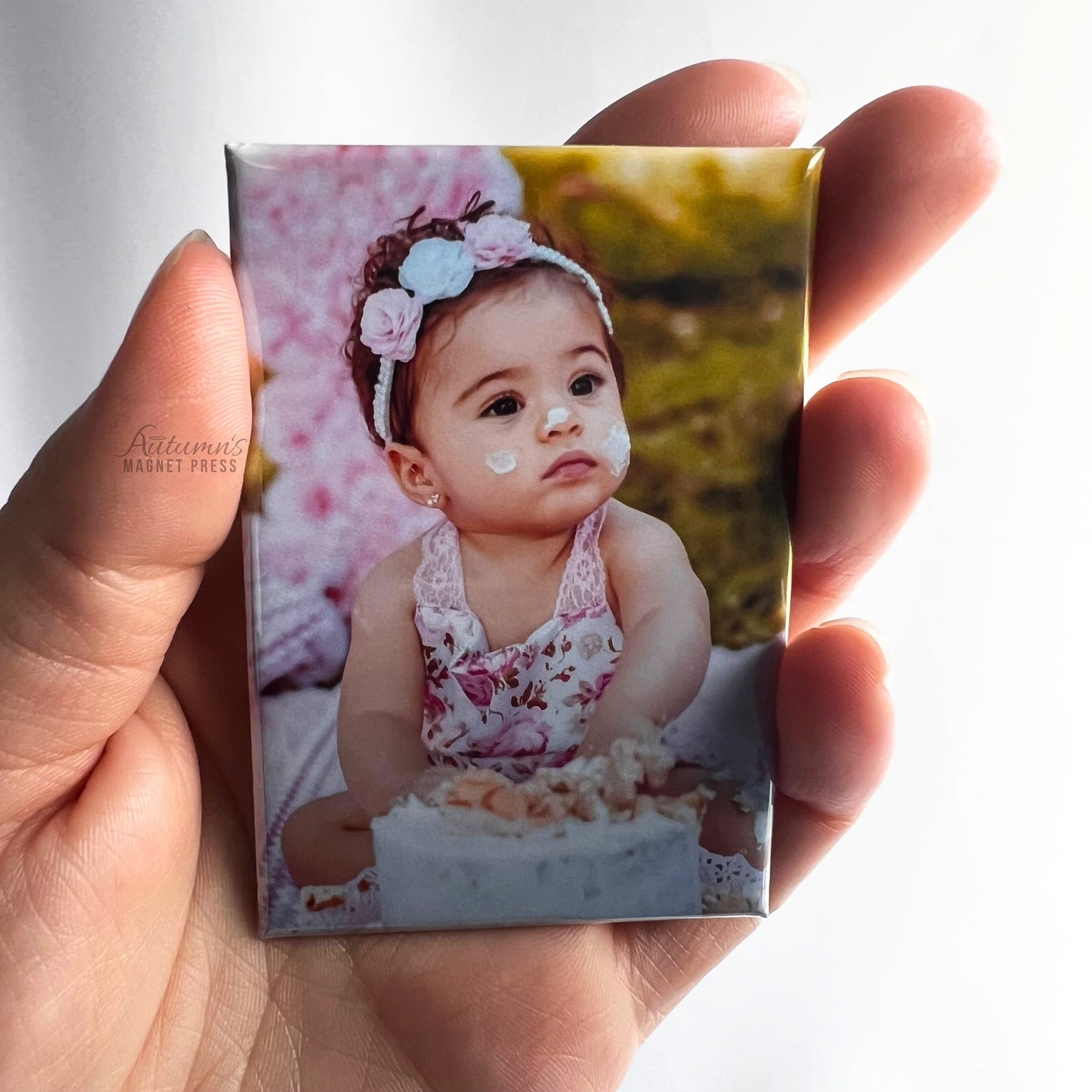 Hand holding a custom photo magnet featuring a baby girl sitting beside her birthday cake during a first birthday celebration against a soft white background.