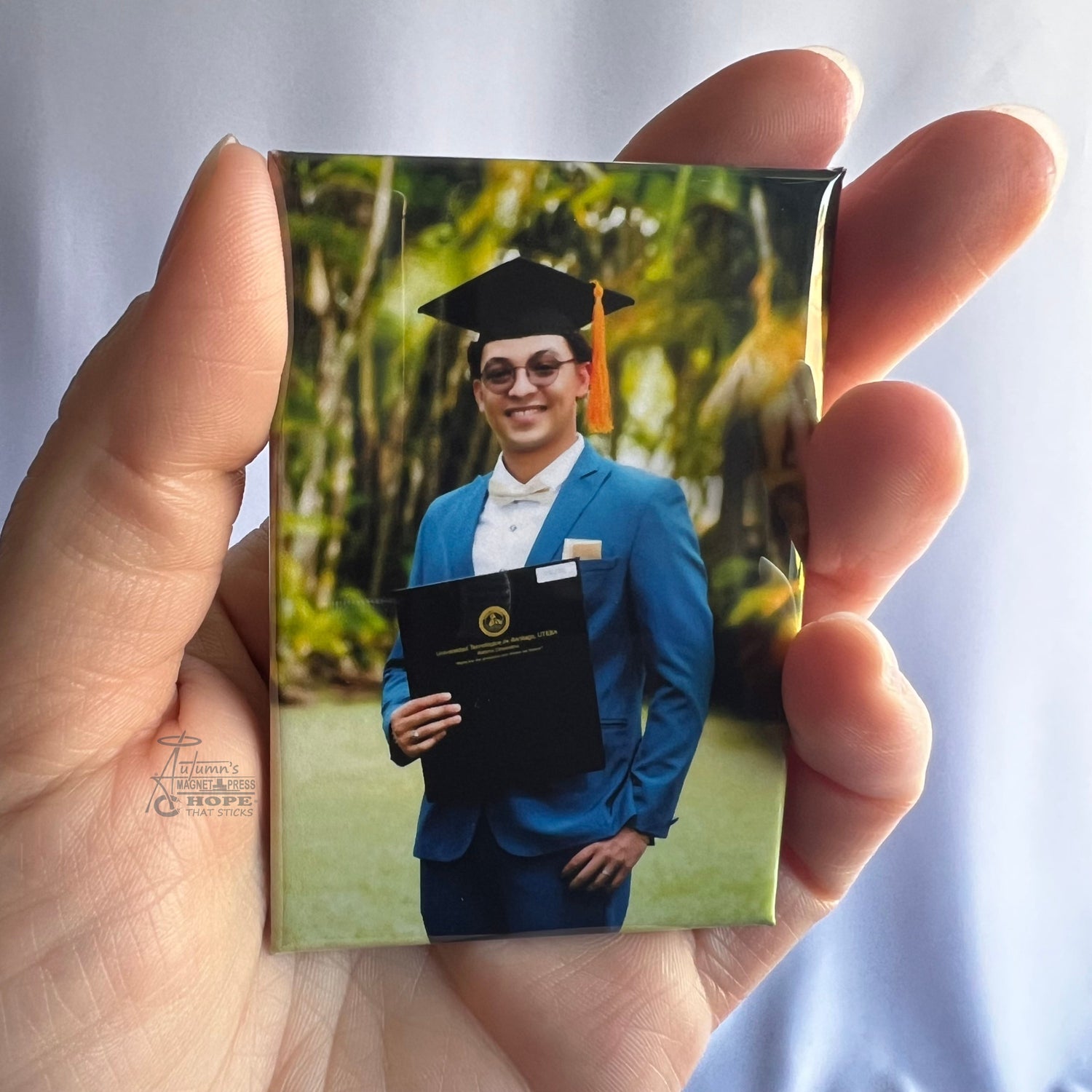 Hand holding a custom graduation photo magnet featuring a graduate in blue cap and gown holding a diploma outdoors against a soft white background