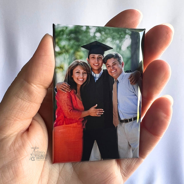 Hand holding a custom graduation photo magnet featuring a graduate posing with proud family members outdoors against a soft white background.
