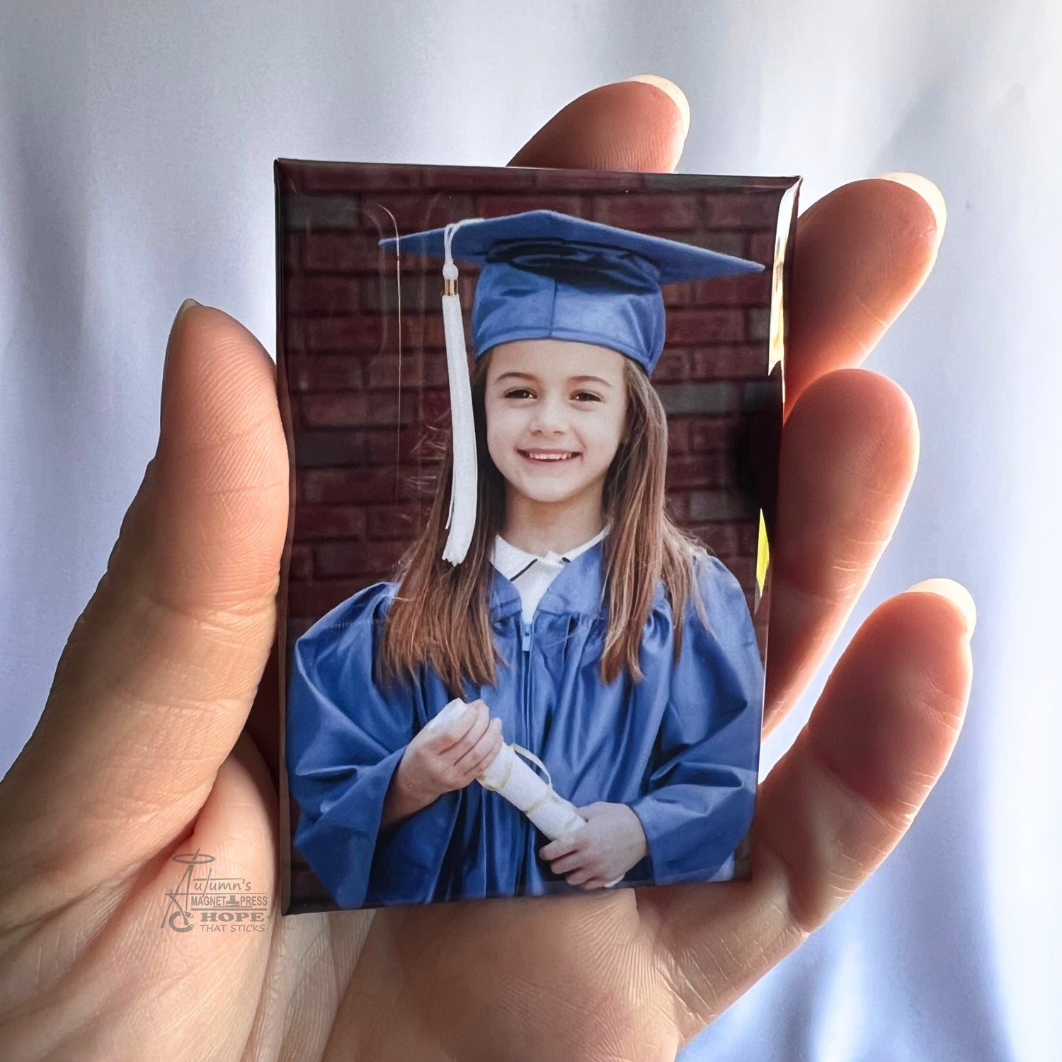 Hand holding a custom kindergarten graduation photo magnet featuring a young girl in blue cap and gown against a soft white background