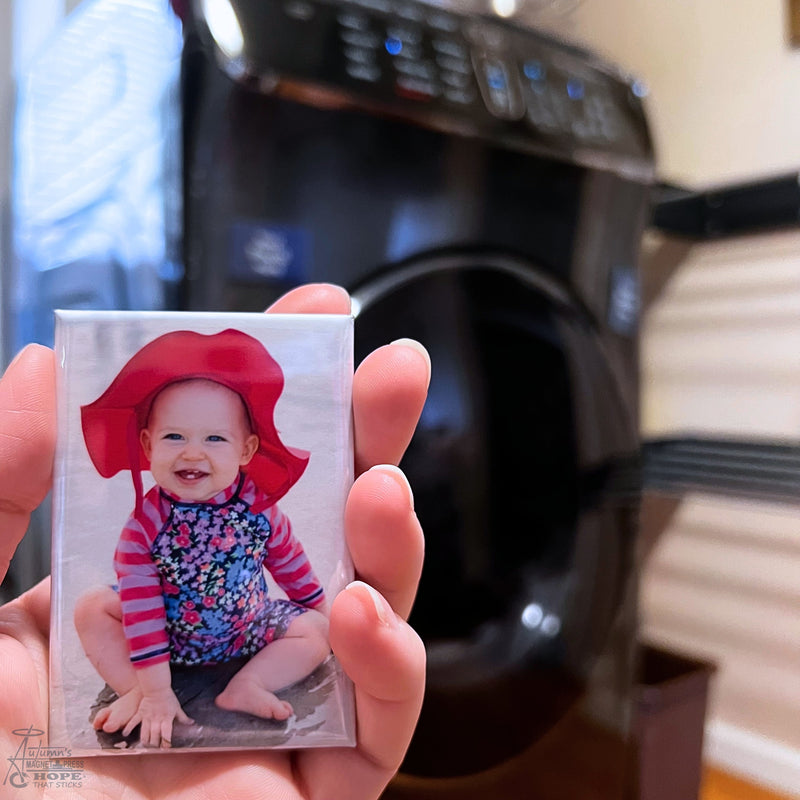 Hand holding a custom baby photo magnet in a laundry room with washing machine blurred in the background