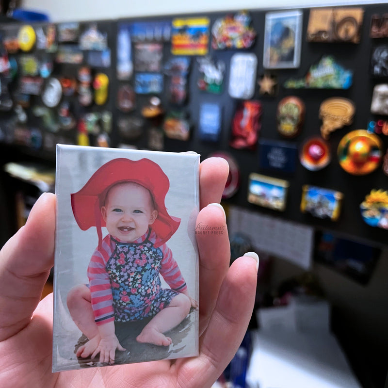 Custom baby photo magnet featuring a smiling baby in a red sun hat held in hand in front of a magnet collection display wall.