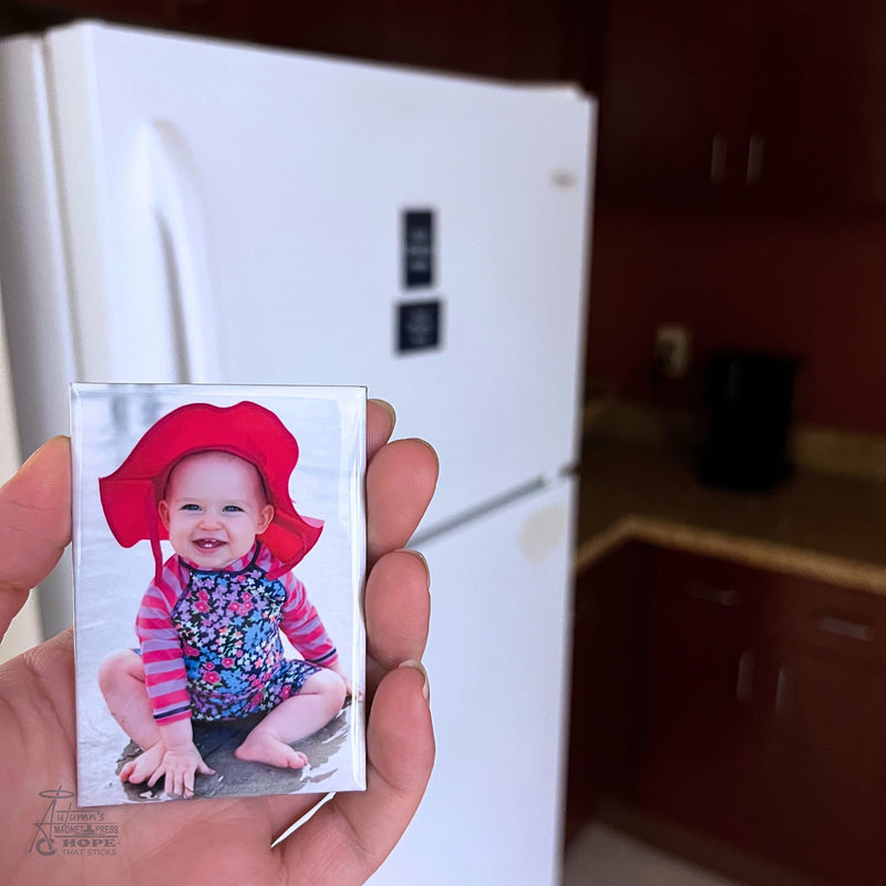 Custom baby photo magnet featuring a smiling baby in a red sun hat held in hand in front of a white refrigerator in a kitchen setting.