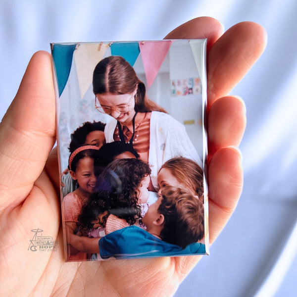 Hand holding a custom teacher appreciation photo magnet featuring a teacher surrounded by smiling students in a classroom against a soft white background
