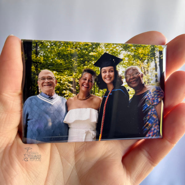 Hand holding a custom graduation photo magnet featuring a graduate posing with parents and grandparent outdoors against a soft white background