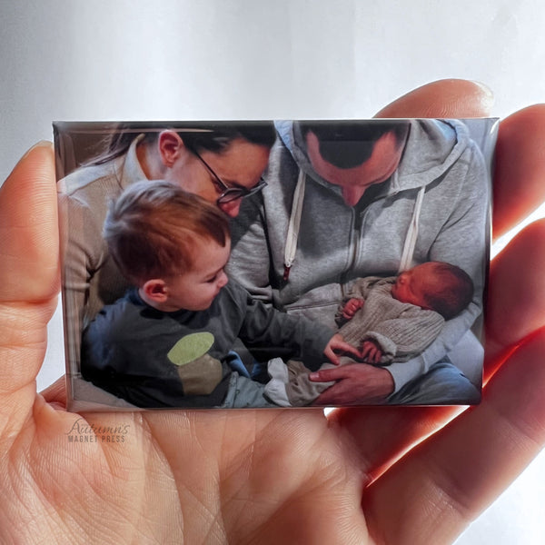 Hand holding a custom photo magnet featuring a toddler meeting and gently touching a newborn sibling for the first time against a soft white background.